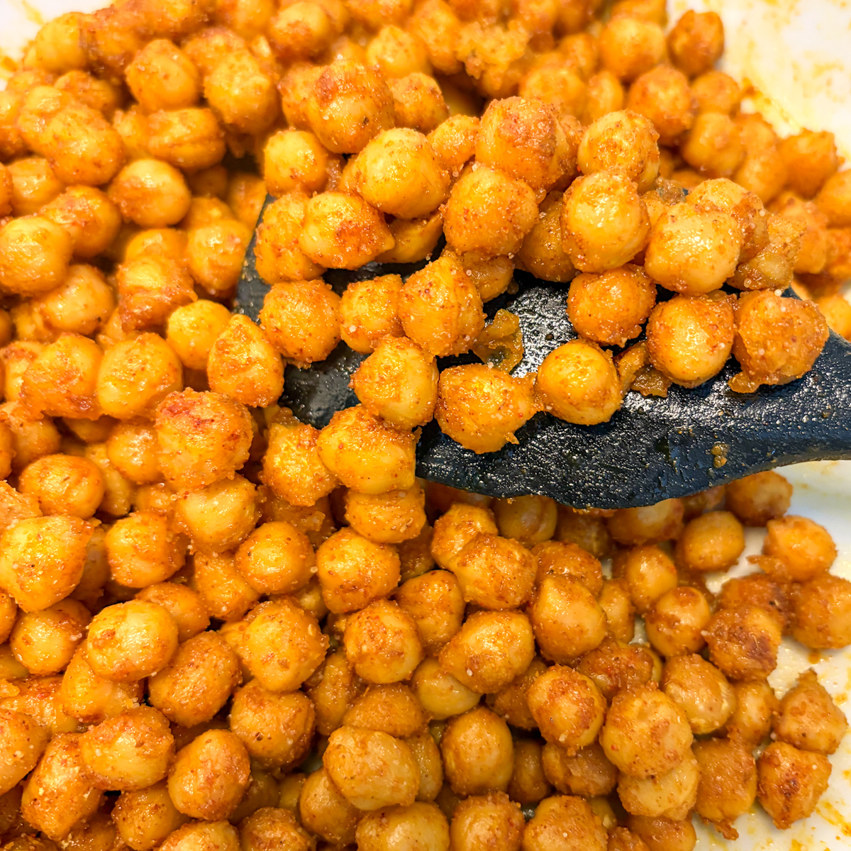 A black spatula scooping up warm, freshly seasoned spiced chickpeas straight from the mixing bowl.