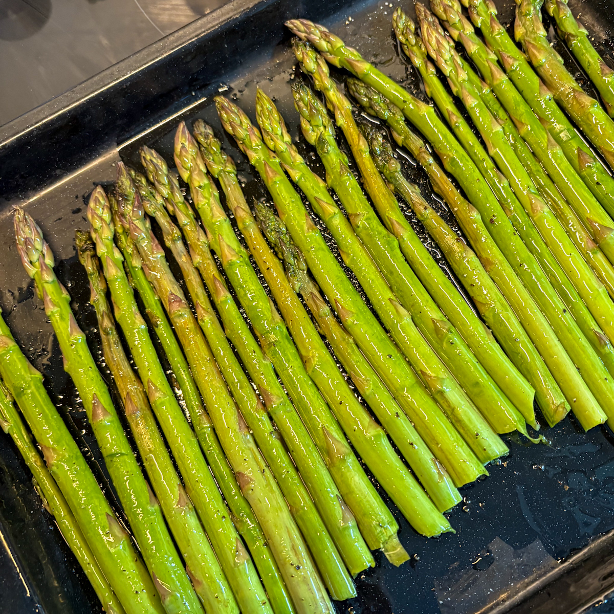 Fresh asparagus drizzled with olive oil on a stainless steel roasting tray, ready for the oven.