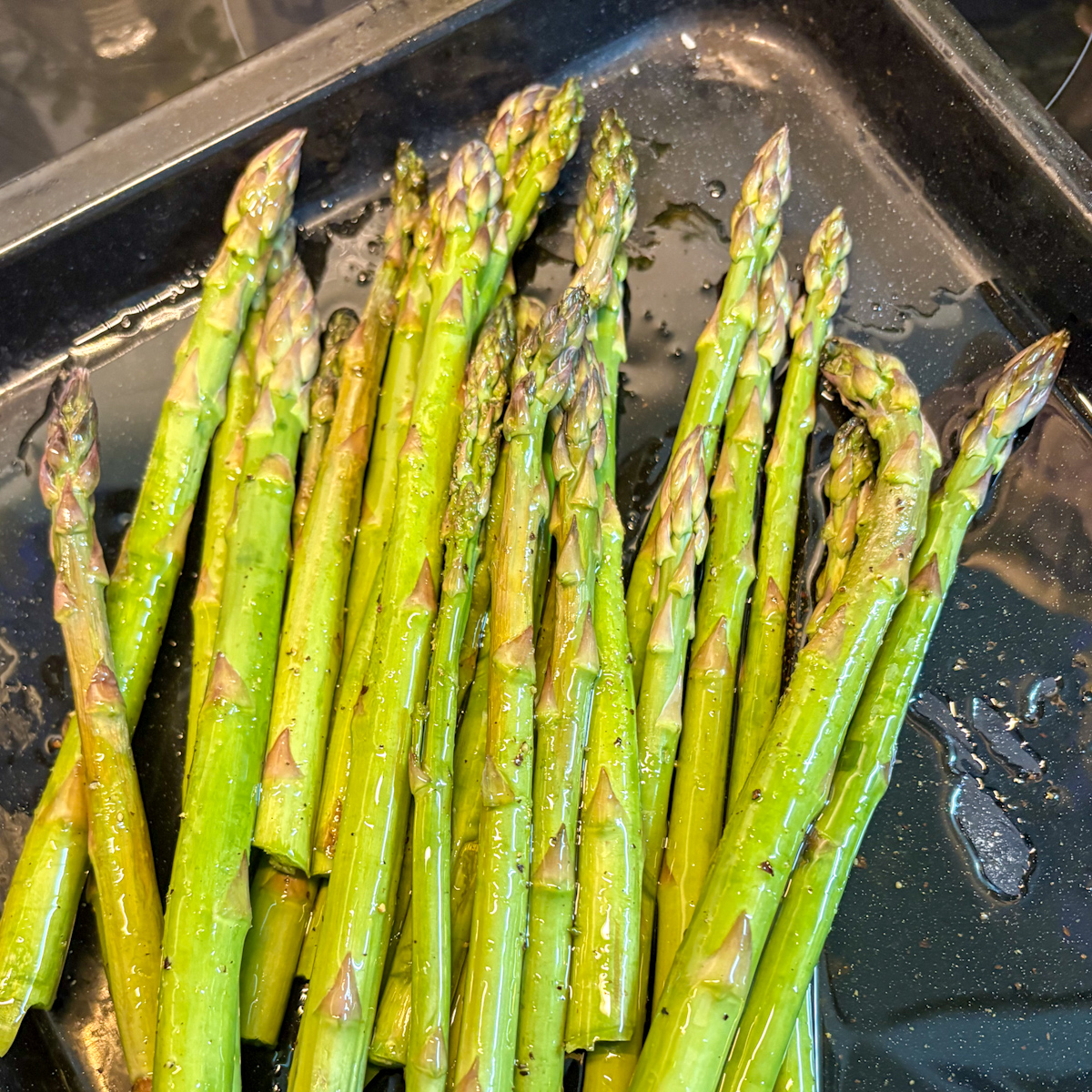 Close-up of raw asparagus spears coated in olive oil on a metal roasting tray before roasting.
