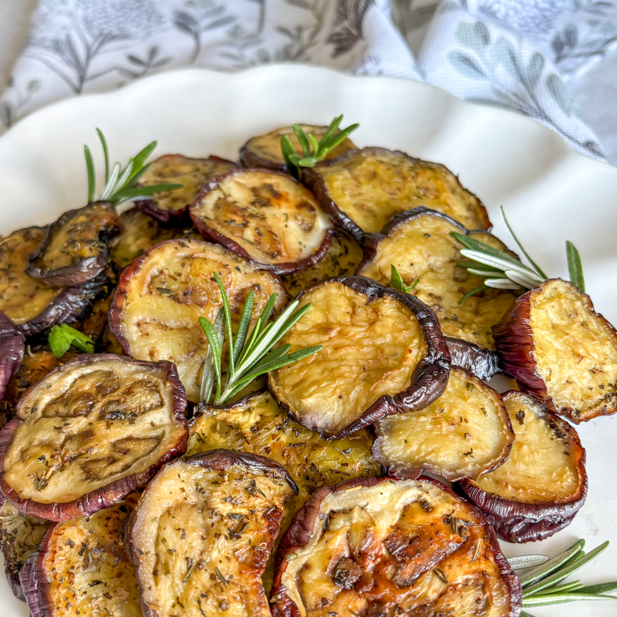 Oven roasted aubergine slices on scalloped plate with herb-print linen