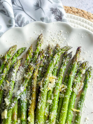 Pan-steamed asparagus on white plate topped with grated Parmesan, lemon zest and garlic butter sauce.