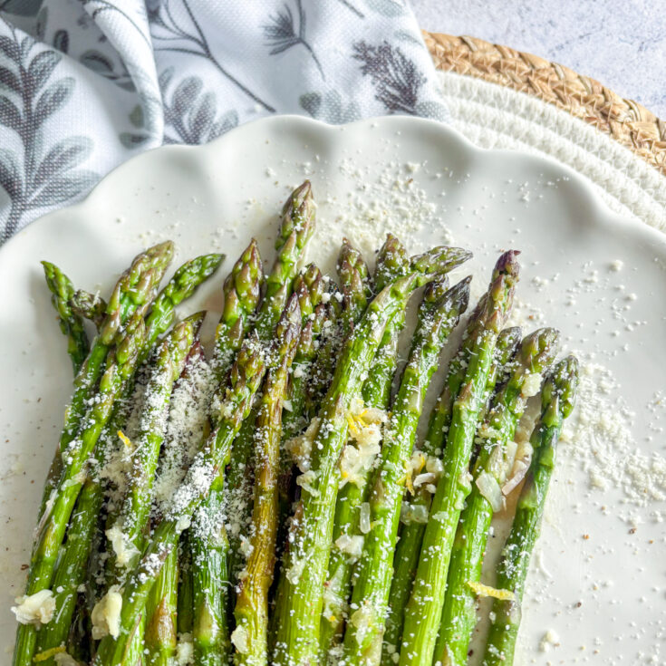 Pan-steamed asparagus on white plate topped with grated Parmesan, lemon zest and garlic butter sauce.