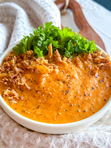 Homemade roasted carrot and ginger soup served in a white bowl with parsley and crispy onions.