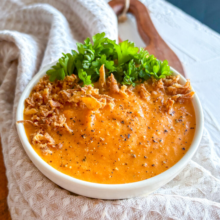 Homemade roasted carrot and ginger soup served in a white bowl with parsley and crispy onions.