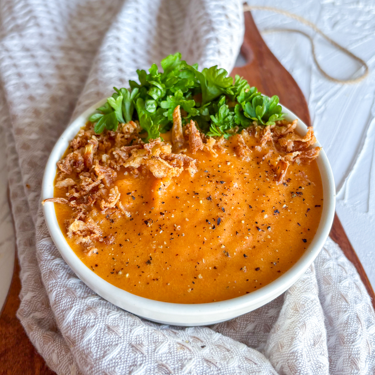 Silky roasted carrot and ginger soup with crispy onions and fresh parsley on a beige tea towel.