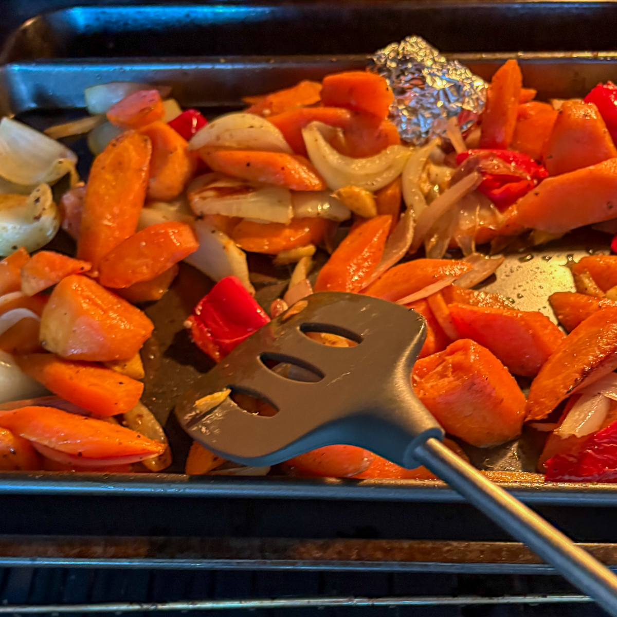Stirring roasted vegetables in the oven for roasted carrot and ginger soup with a black spatula.