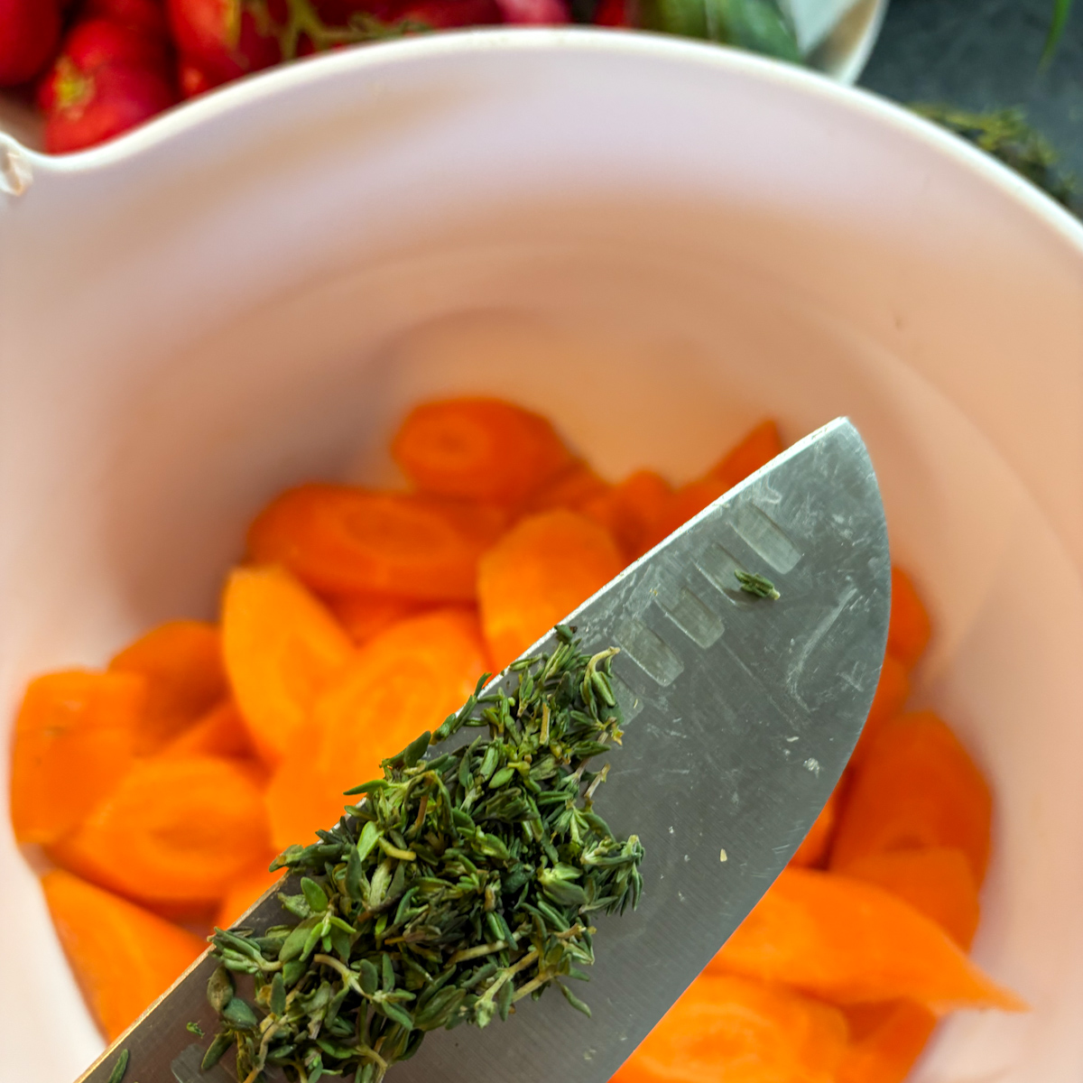 Knife with minced fresh thyme leaves over sliced peeled carrots in a pink mixing bowl