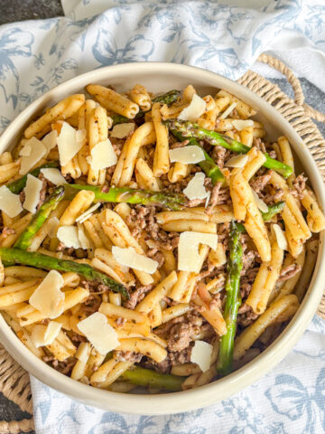 Beef and asparagus pasta in a white bowl with shaved parmesan and crunchy asparagus tips on a blue floral napkin