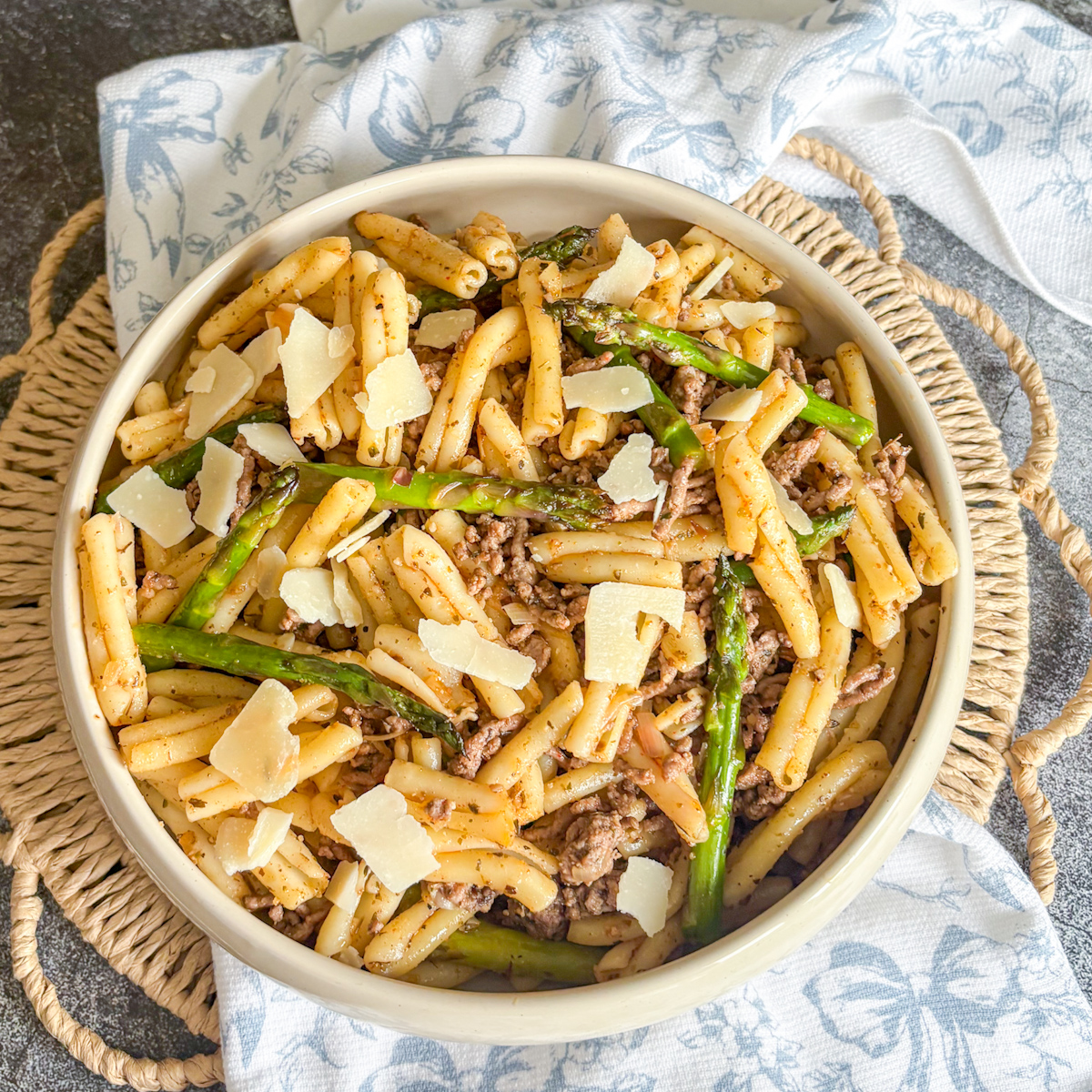 Beef and asparagus pasta in a white bowl with shaved parmesan and crunchy asparagus tips on a blue floral napkin