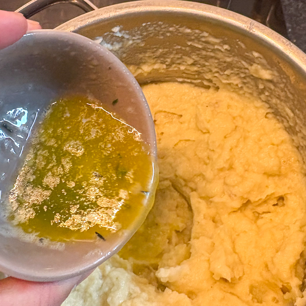 Pouring melted thyme butter from a small bowl into a pot of parsnip and pear mash being prepared.
