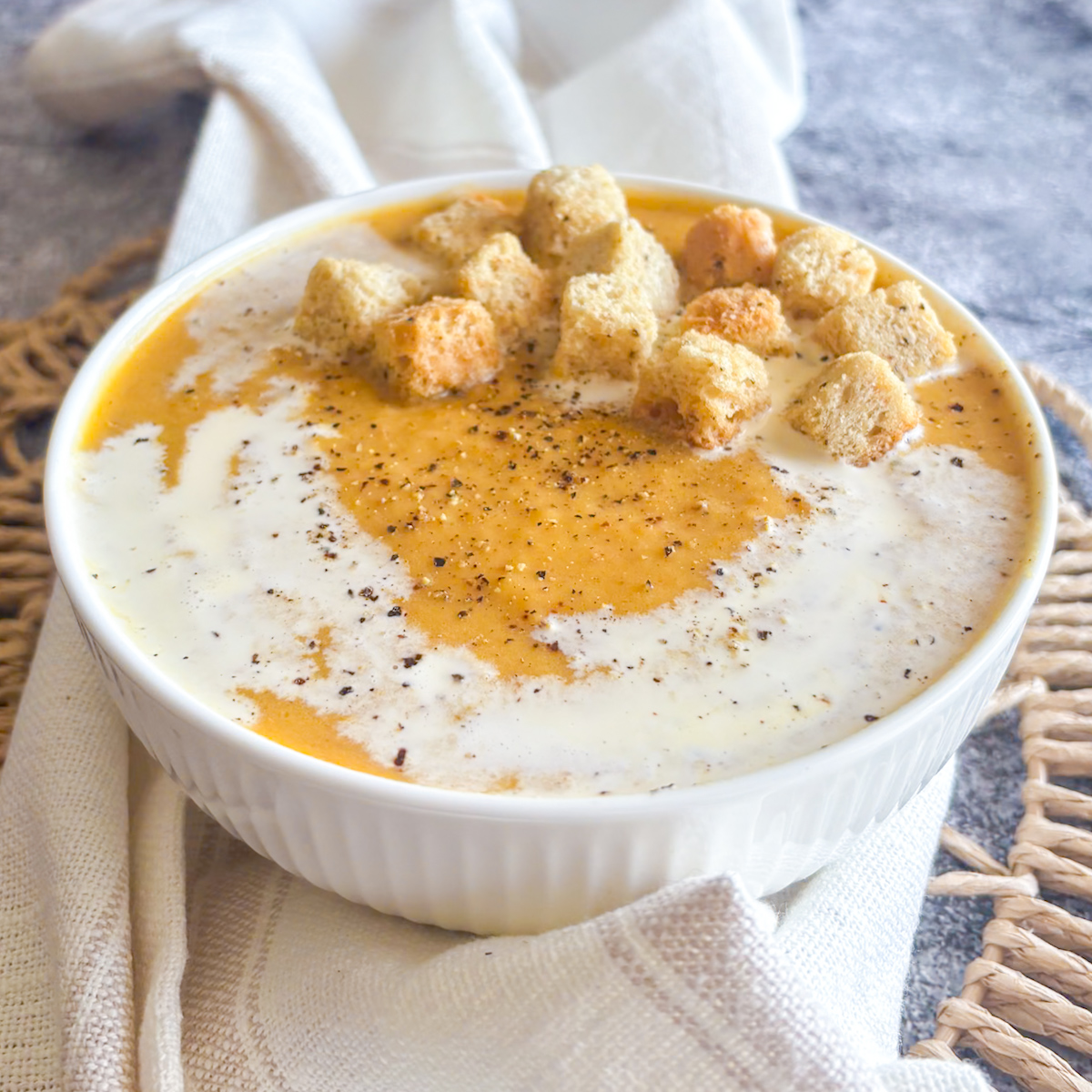 Creamy roasted root vegetable soup in white bowl with croutons, cream swirl and cracked black pepper on top