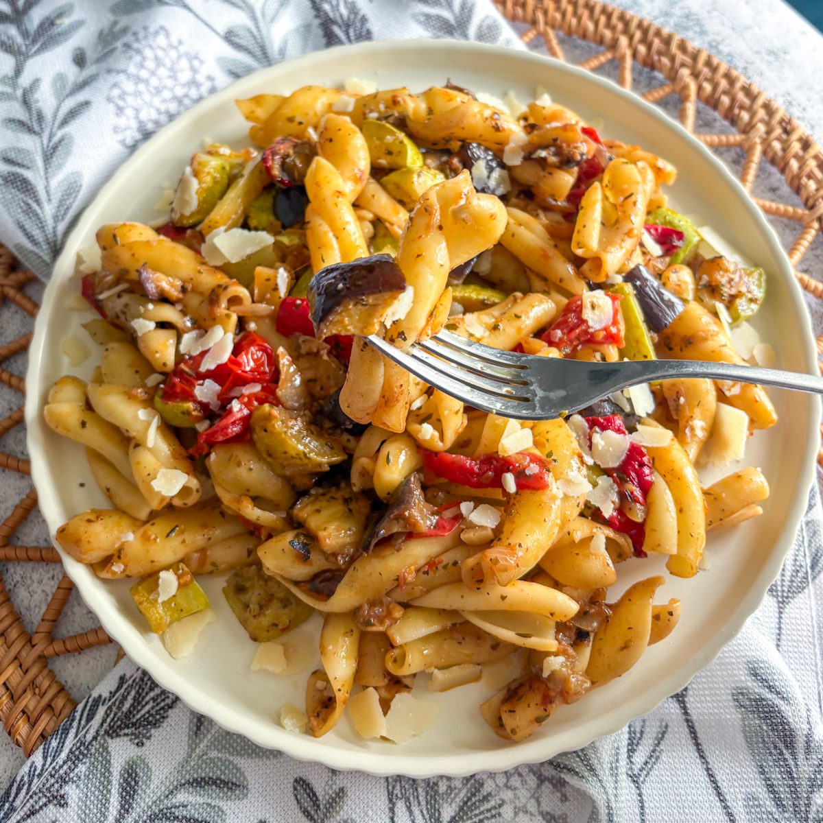 Fork lifting roasted vegetable pasta with aubergine, courgette and cherry tomatoes from the plate