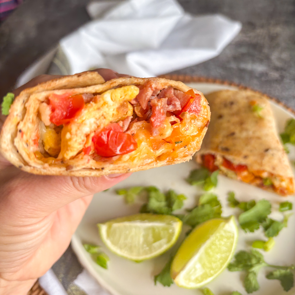 Hand holding half a breakfast burrito with bacon, egg and tomato inside