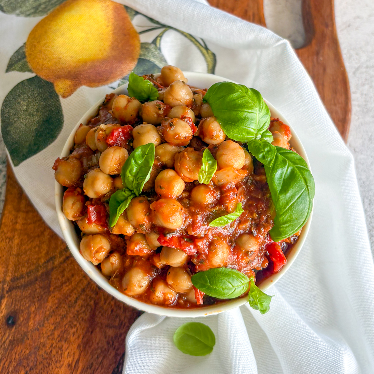Overhead bowl of chickpeas in tomato sauce with basil and a lemon napkin