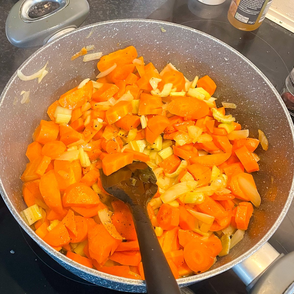 Golden sautéed carrots, onion and celery in a grey pan with a black spatula, base for chilled carrot soup recipe.
