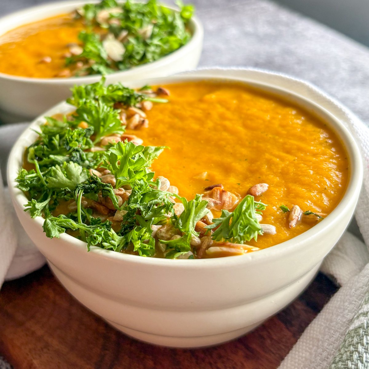  Bright chilled carrot soup in white bowls with parsley, almond flakes and sunflower seeds on a wooden board.