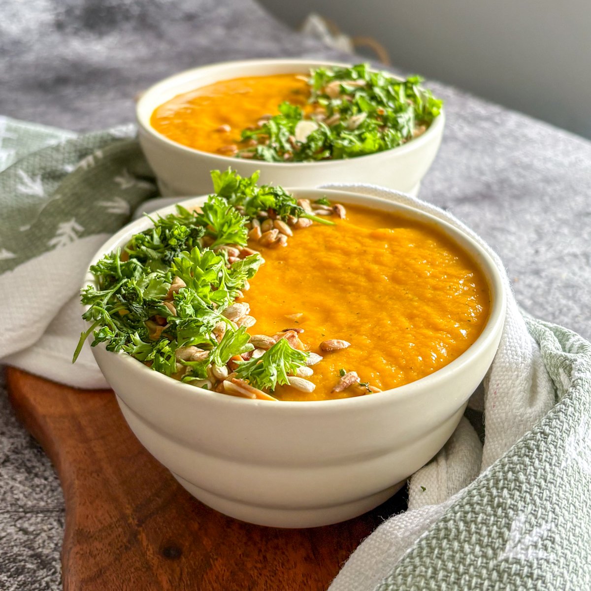 Vibrant chilled carrot soup overhead in two bowls with fresh parsley, sunflower seeds and a linen napkin.