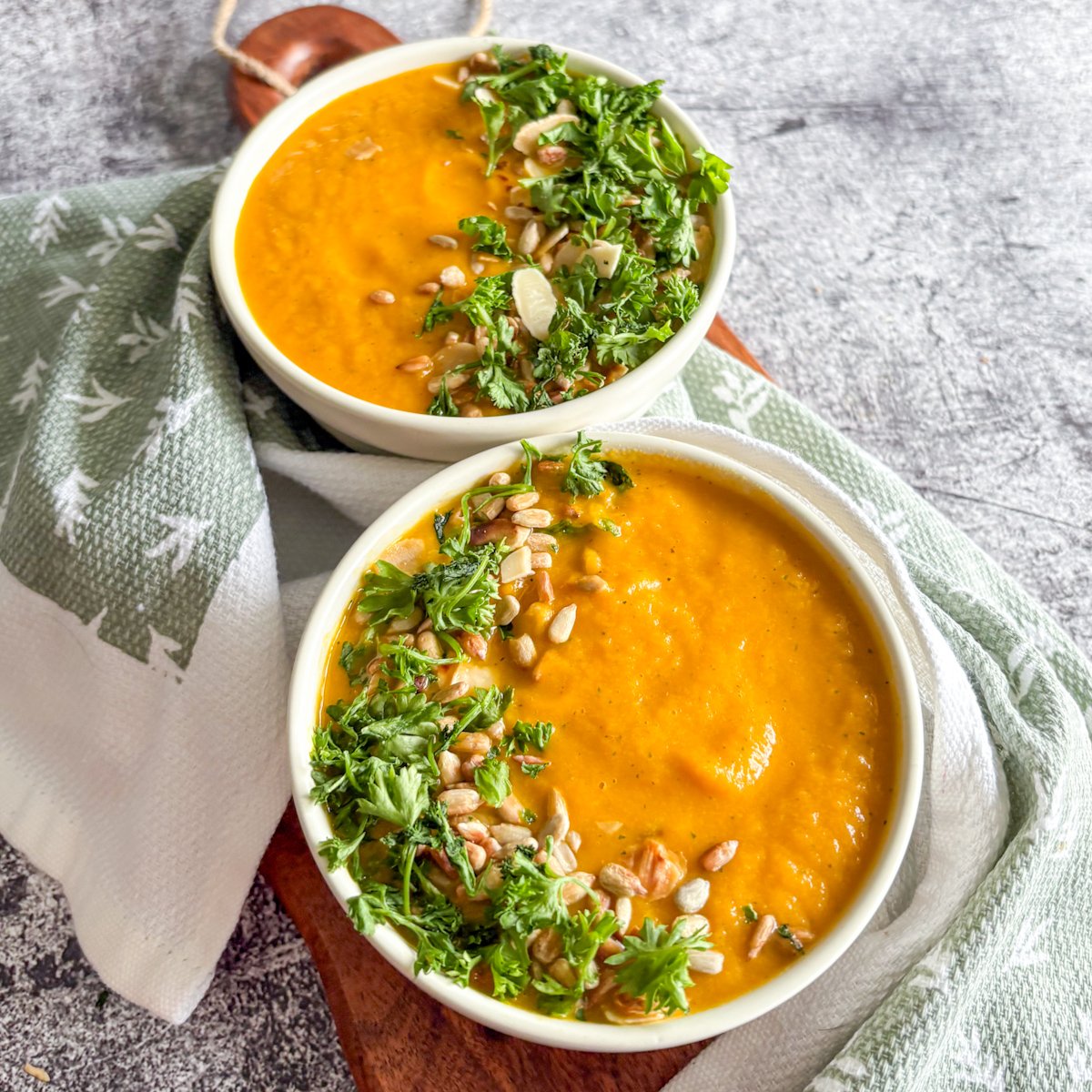 Silky chilled carrot soup in two white bowls topped with curly parsley, almond flakes and toasted sunflower seeds.