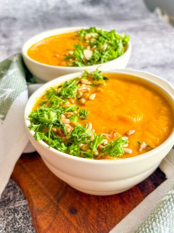 Refreshing chilled carrot soup served in white bowls with parsley, almond flakes and sunflower seeds on a wooden board.