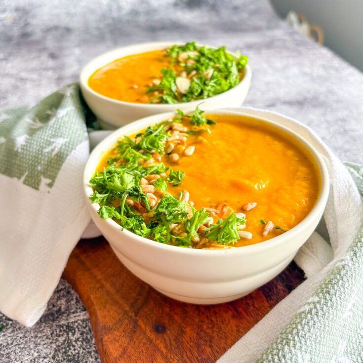 Refreshing chilled carrot soup served in white bowls with parsley, almond flakes and sunflower seeds on a wooden board.
