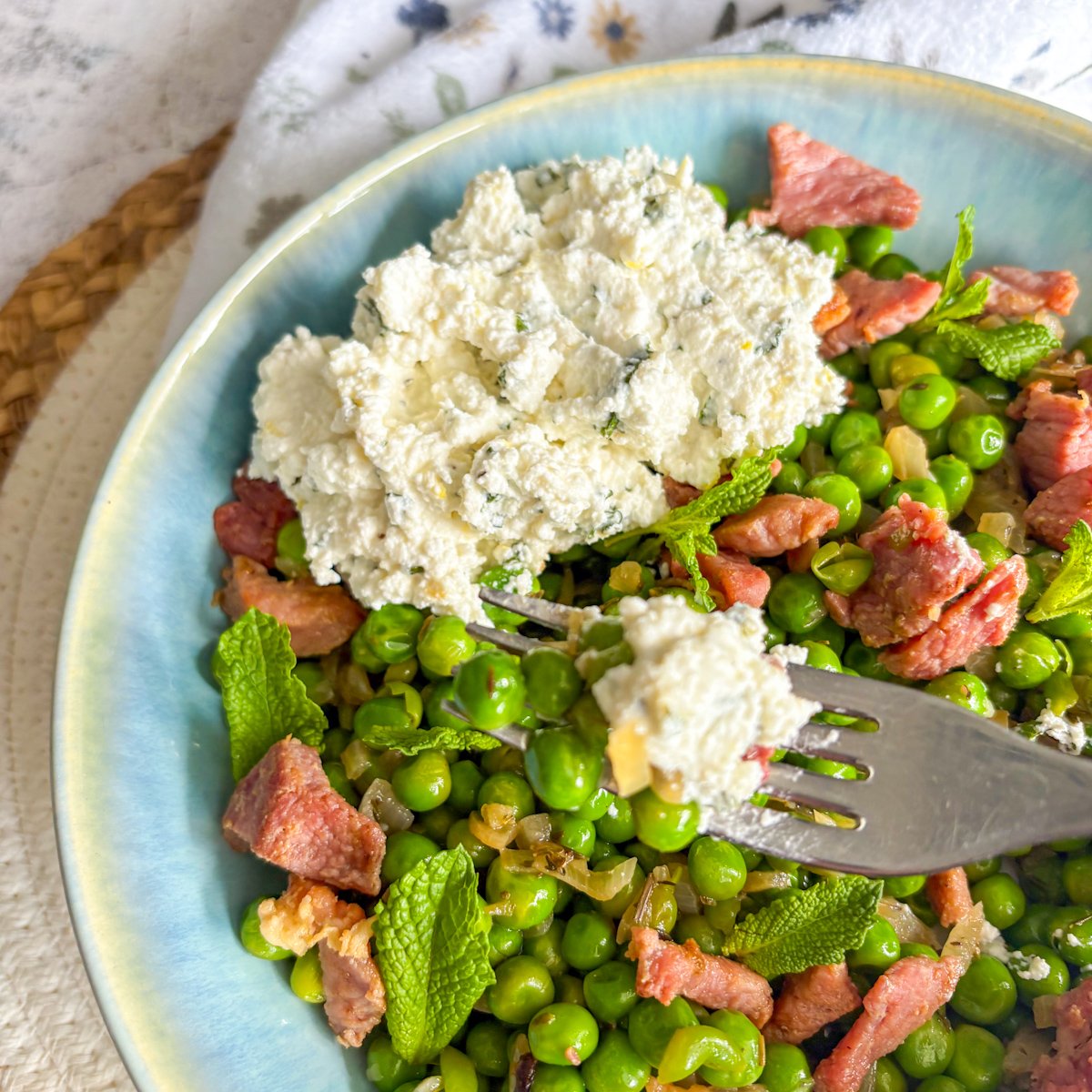 Fork lifting peas with bacon and ricotta from a blue bowl with fresh mint