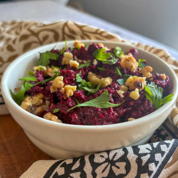 Beetroot and walnut dip in a sunlit white bowl on a wooden board, topped with crunchy walnuts and parsley