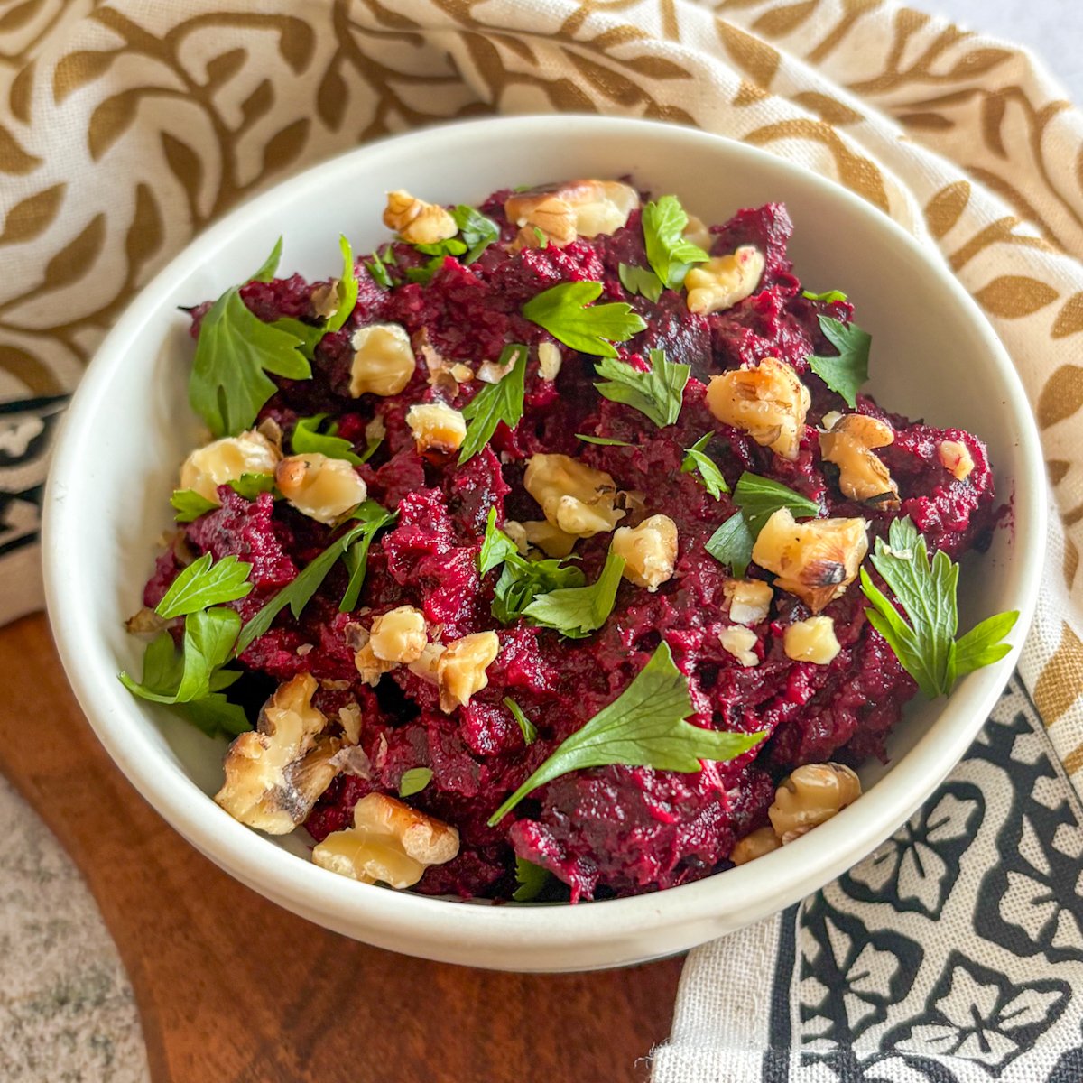 Beetroot and walnut dip in a cream bowl framed by gold leaf-print and black patterned tea towels on a board