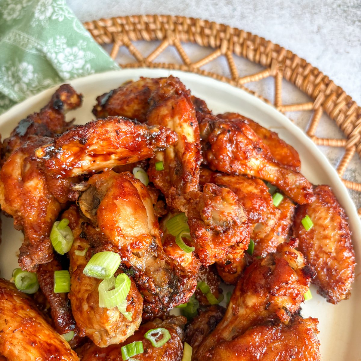 Guinness chicken wings on a cream plate beside a woven mat and green floral napkin, deeply glazed and sprinkled with spring onions.