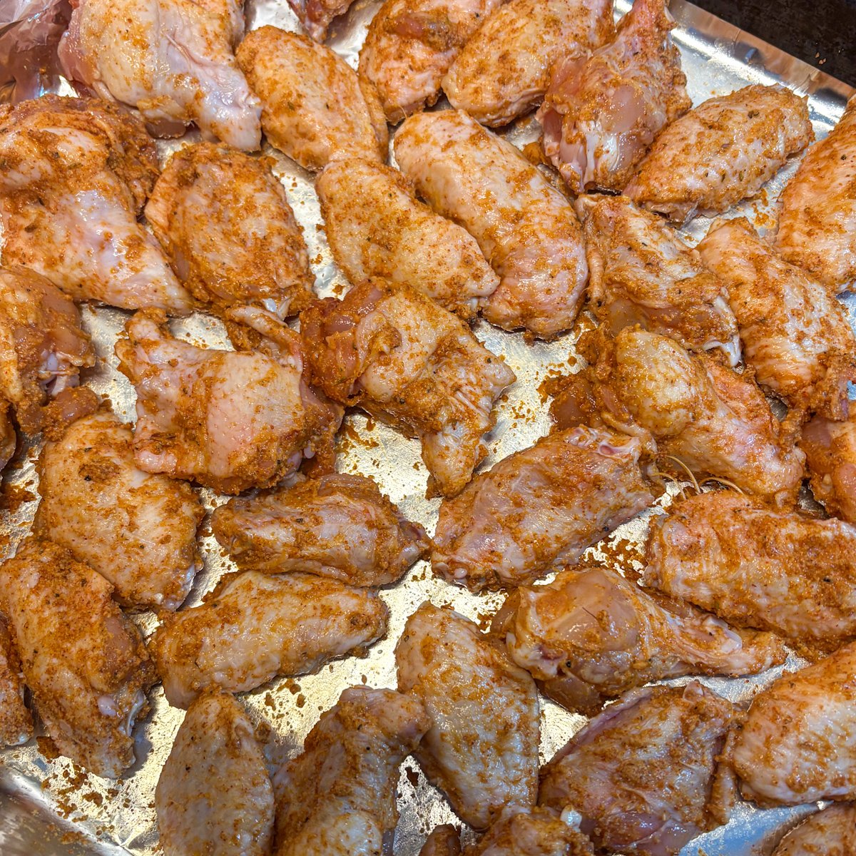 Raw chicken wings tossed in a smoky red dry rub, spread out on a foil-lined baking tray before baking.