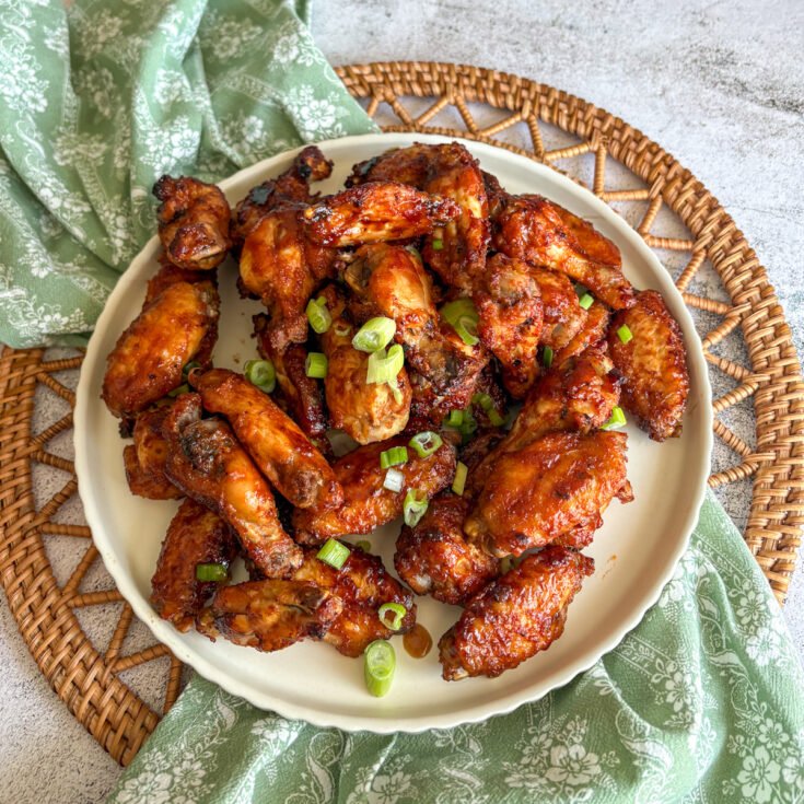 Baked Guinness chicken wings piled on a plate with a green floral napkin and woven mat, glazed and topped with spring onions.