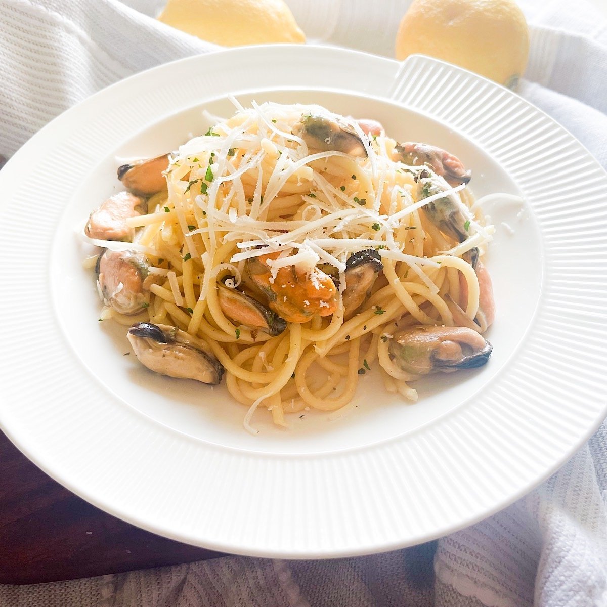 Mussel pasta in creamy garlic butter sauce served on a white plate with grated parmesan, parsley and fresh lemons in the background