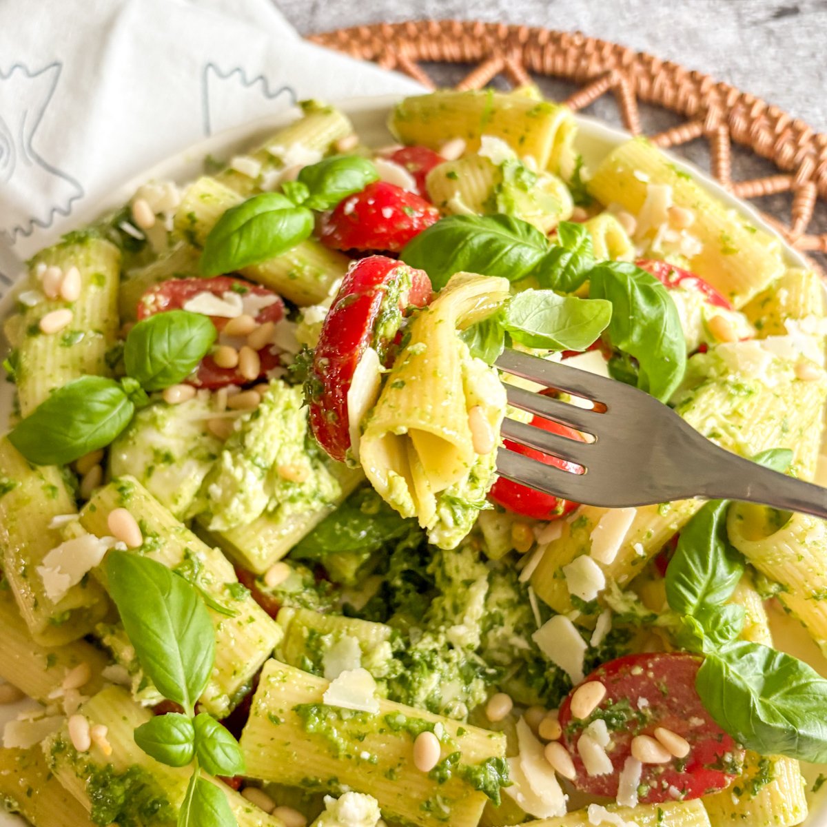 Fork lifting rigatoni and tomato from a summer pesto pasta salad with basil and pine nuts