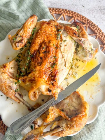 Overhead view of herb butter roast chicken being carved, one breast sliced open to show the herb butter layer under the skin, with a knife, thyme sprigs and buttery juices on the plate.