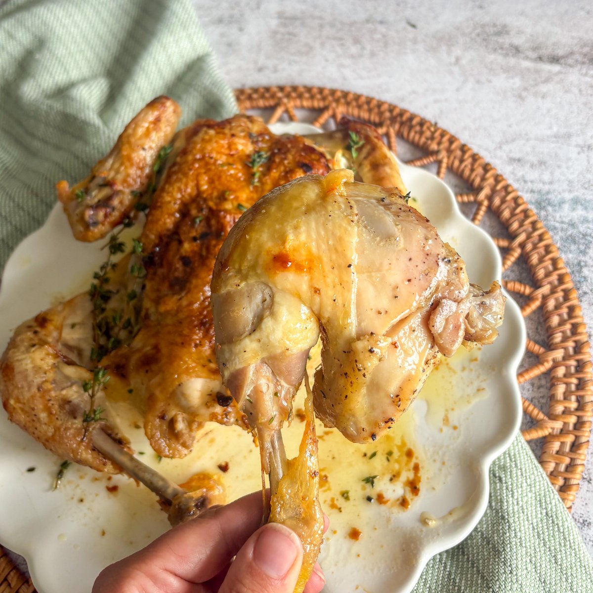 A hand lifting a whole roasted chicken leg away from the herb butter roast chicken, showing the tender meat and buttery juices left behind on the white scalloped plate.