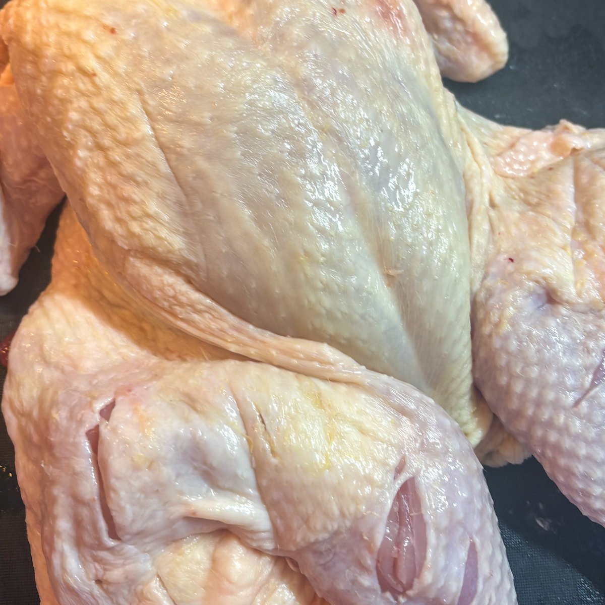Raw spatchcocked chicken lying flat on a dark board, backbone removed and breastbone pressed down, with shallow slashes cut into the thighs and legs ready for brining before making herb butter roast chicken.