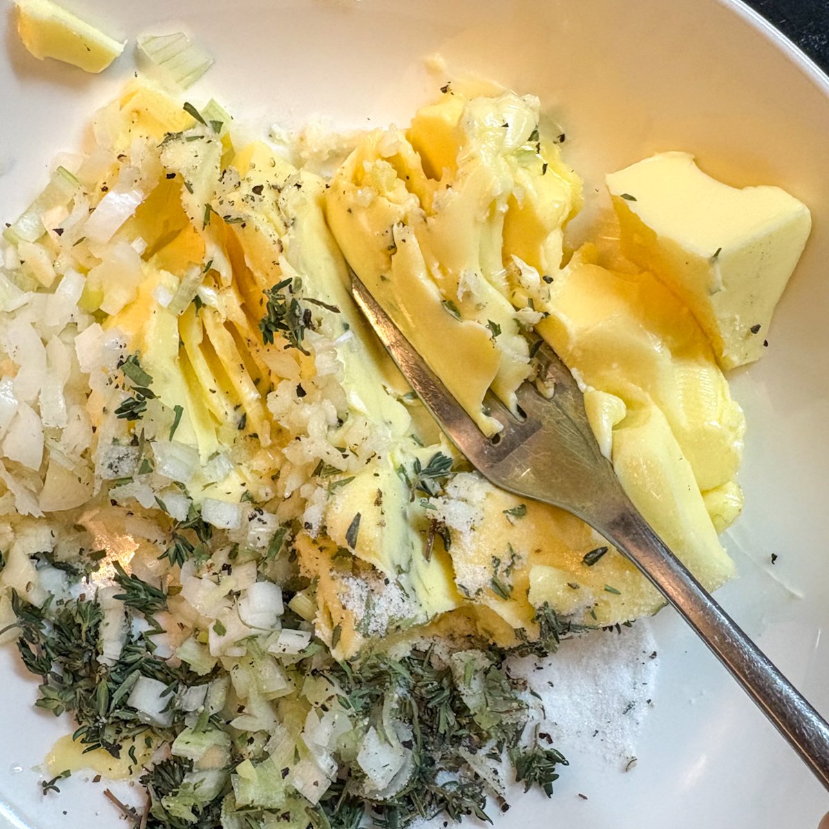 Herb butter for roast chicken being mashed together with a fork in a white bowl — softened yellow butter, finely chopped spring onion whites, minced garlic, dried tarragon, fresh thyme leaves, salt and cracked black pepper.