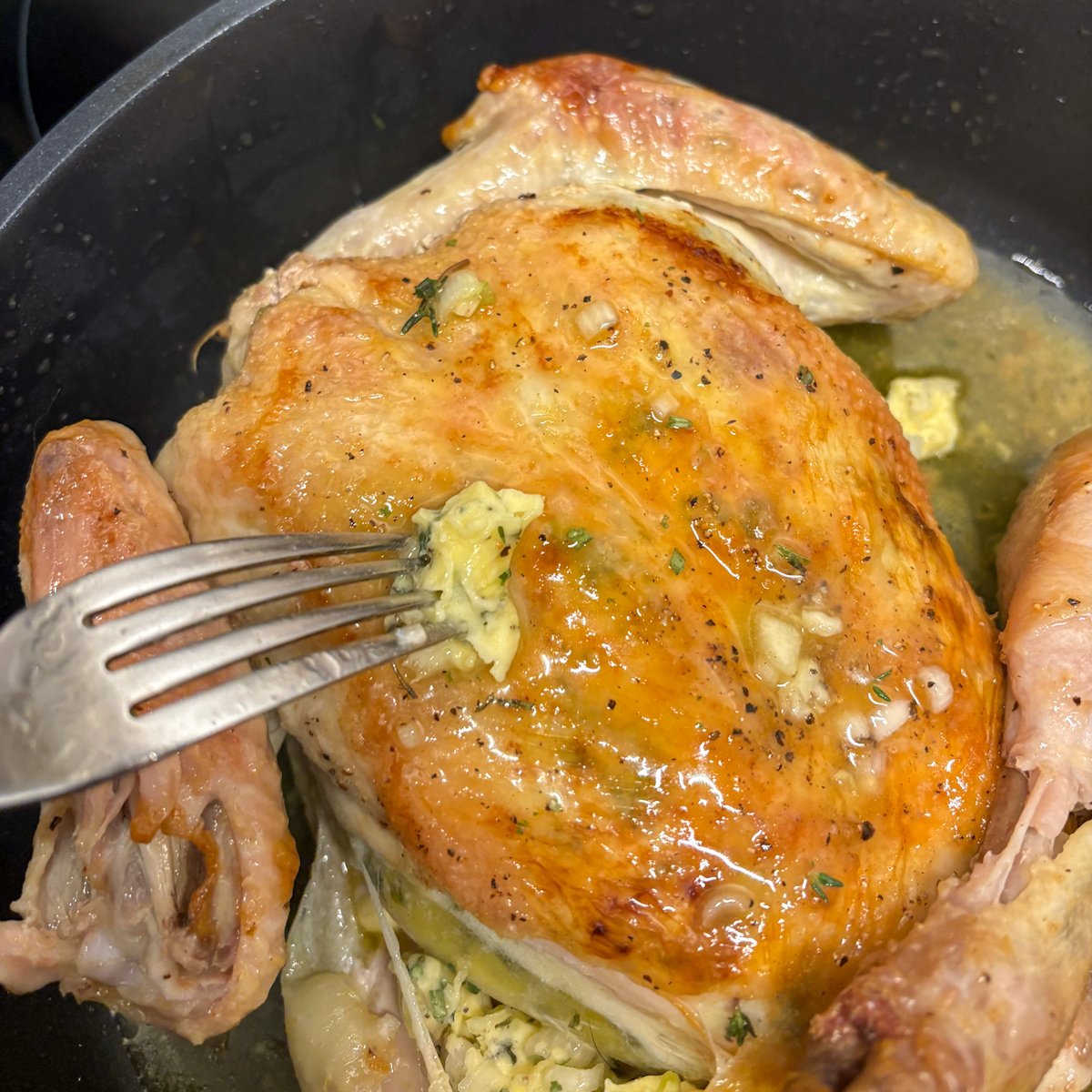 Part-roasted herb butter roast chicken in a dark skillet, skin side up and golden, being coated with the reserved herb butter using a fork before returning to the oven.