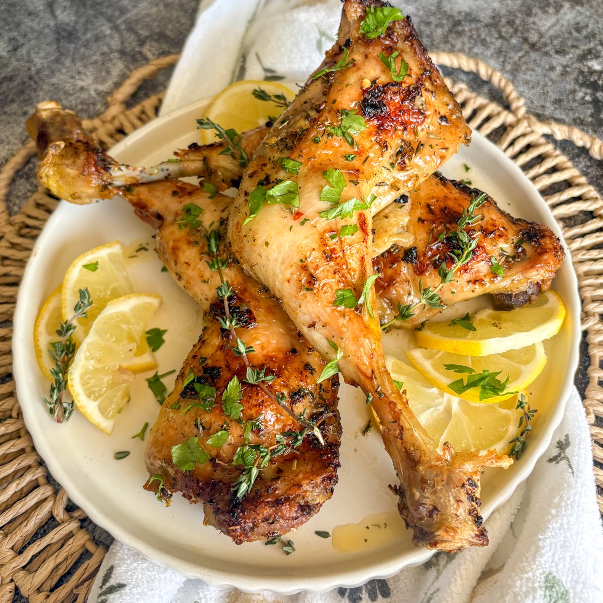Baked lemon chicken legs arranged with the drumstick bones pointing upward, dressed with lemon slices and fresh parsley, photographed from above on a cream plate over a woven basket, showing the herb-flecked, golden skin of this budget-friendly oven chicken recipe.