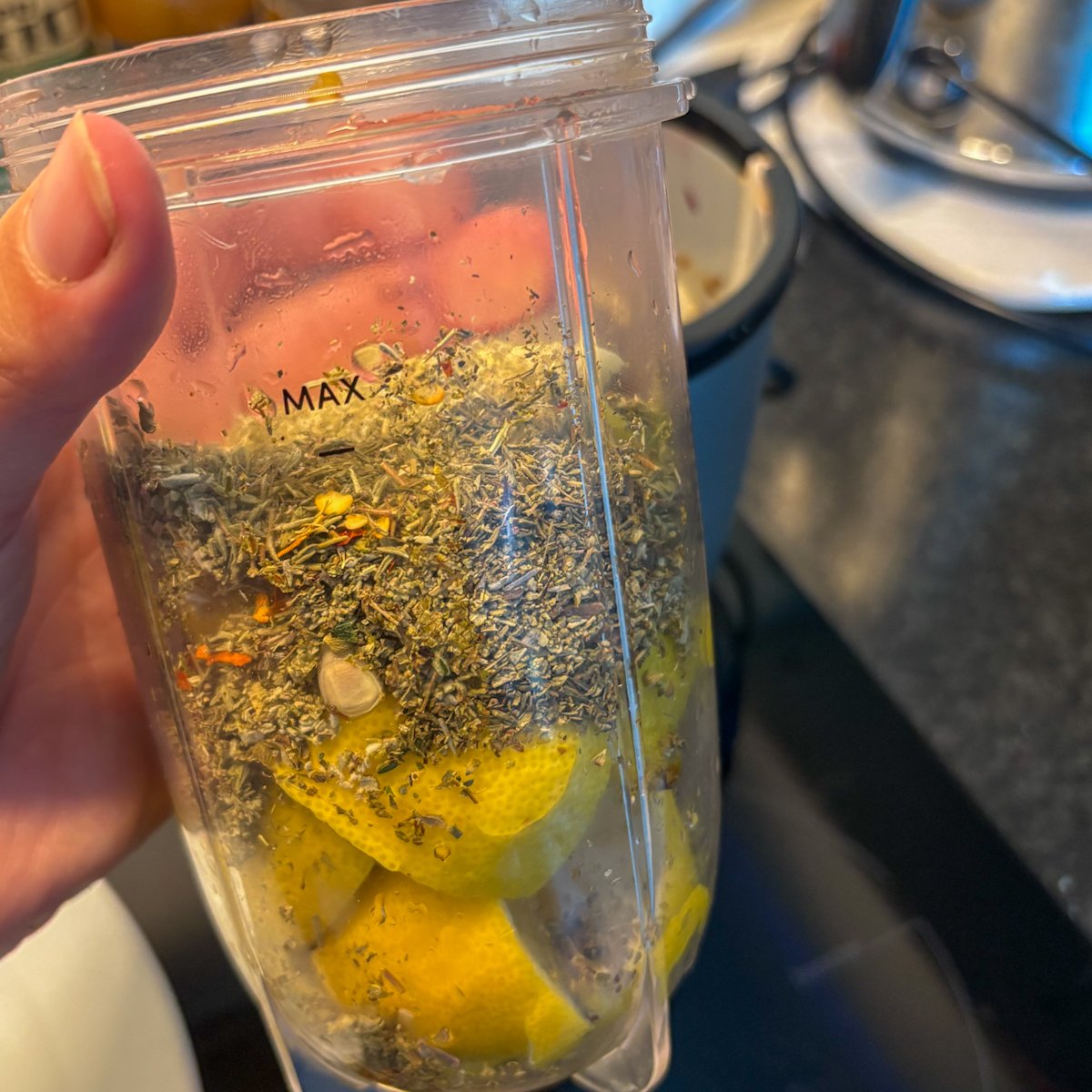 A hand holding a clear blender jug filled with lemon chunks and blitzed dried herbs up to the MAX line, kitchen hob in the background, capturing the first step of making the fresh marinade for these easy lemon chicken legs.
