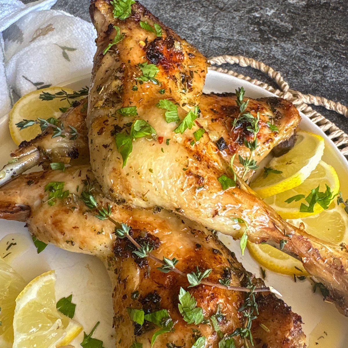 Close, angled shot of glazed lemon chicken legs garnished with fresh parsley and thyme and set beside thin lemon slices against a grey stone background, capturing the juicy dark meat and crisp golden skin of this cheap garlic and herb chicken recipe.