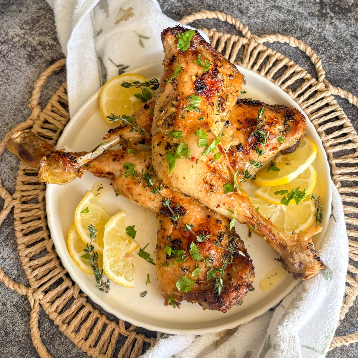  Overhead shot of golden glazed lemon chicken legs on a cream plate with lemon slices and fresh parsley scattered over the top, styled on a woven basket with a floral tea towel, showing the sticky honey butter glaze on this budget family chicken recipe.