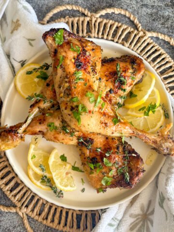 Golden lemon chicken legs stacked on a cream plate with fresh lemon slices arranged around the edge and a scatter of parsley and thyme, set on a woven basket with a white floral tea towel against a grey stone background, showing the crisp glazed skin of this easy budget oven or air fryer chicken dinner.