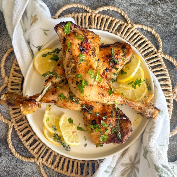 Golden lemon chicken legs stacked on a cream plate with fresh lemon slices arranged around the edge and a scatter of parsley and thyme, set on a woven basket with a white floral tea towel against a grey stone background, showing the crisp glazed skin of this easy budget oven or air fryer chicken dinner.