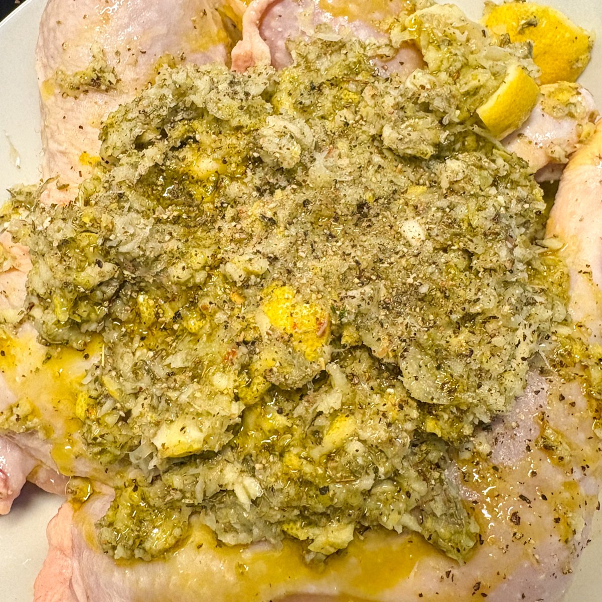 Close-up of a blended green lemon and herb marinade spread thickly over raw chicken legs in a white bowl with a few lemon chunks visible, showing how the fresh garlic and herb marinade coats the meat for these budget lemon chicken legs.