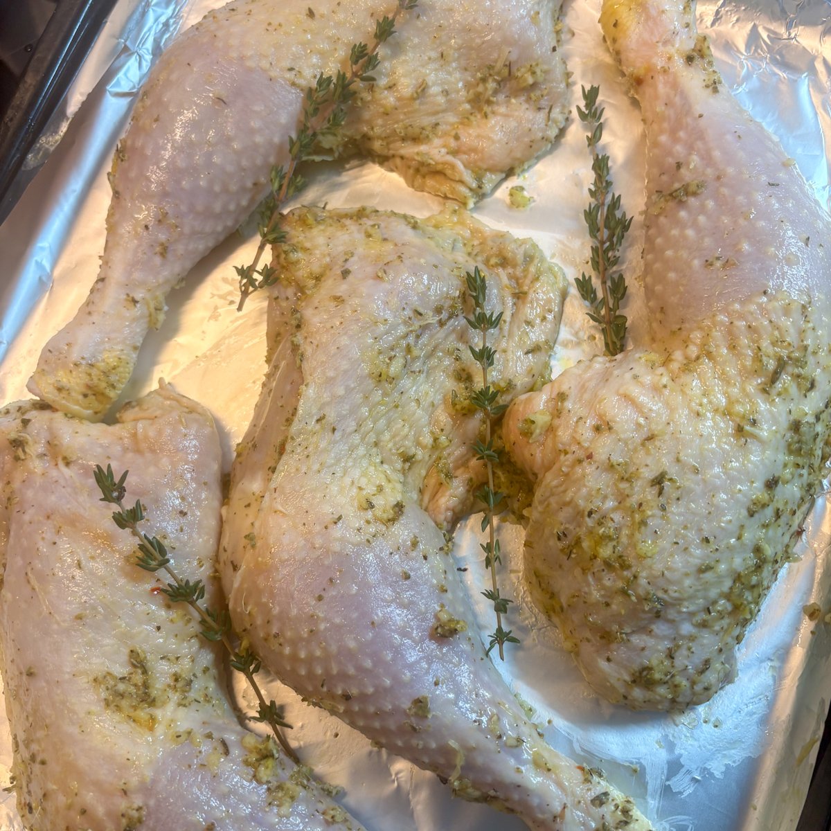 Raw marinated lemon chicken legs laid out on a foil-lined tray with fresh thyme sprigs, coated in a green garlic and herb rub, showing the marinating stage before baking in this easy oven or air fryer chicken recipe.