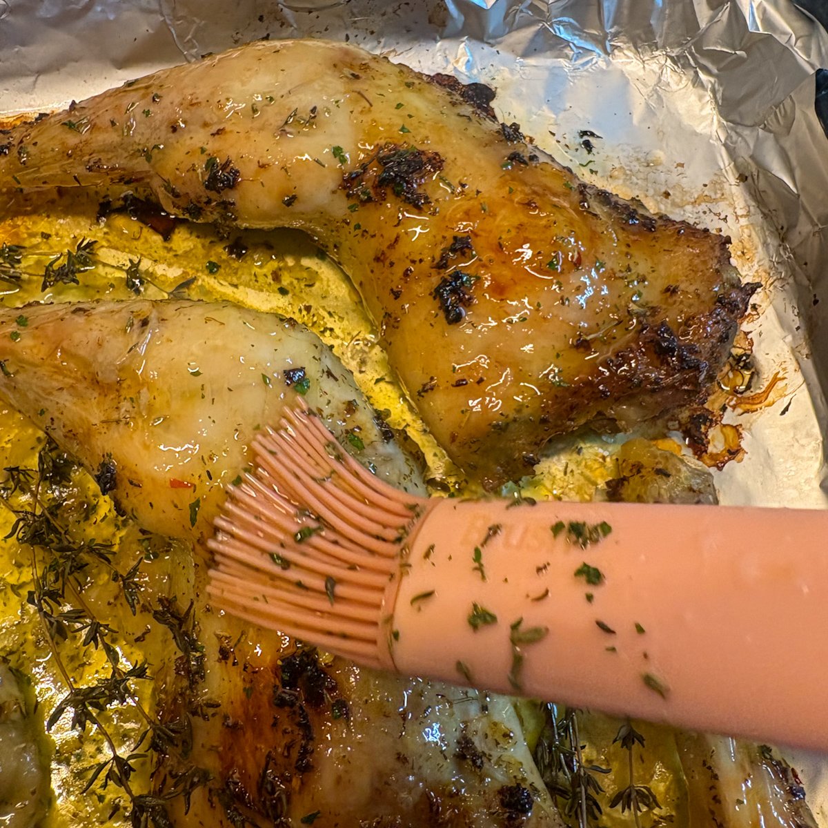 Honey butter glaze being brushed over lemon chicken legs with a pink silicone brush in a foil-lined roasting tray, fresh thyme sprigs tucked around the meat, showing the final basting step that gives this budget chicken recipe its sticky, caramelised finish.