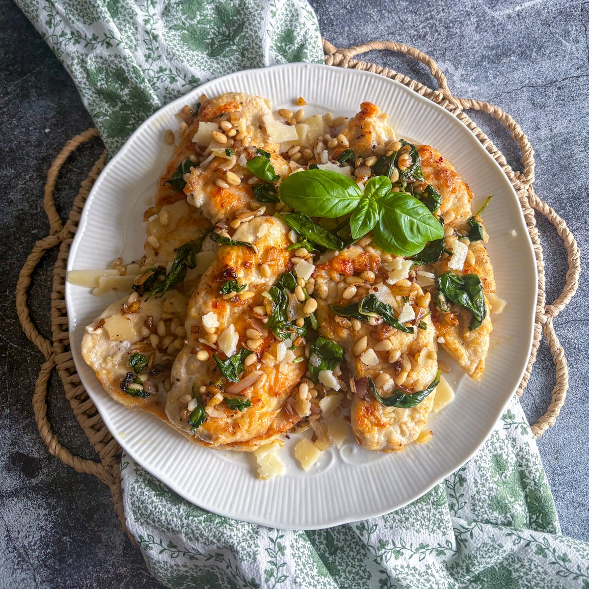 Overhead view of pan-fried chicken cutlets with basil and pine nuts overlapping on a white plate, golden fried edges showing between the wilted basil leaves and Parmesan shavings.