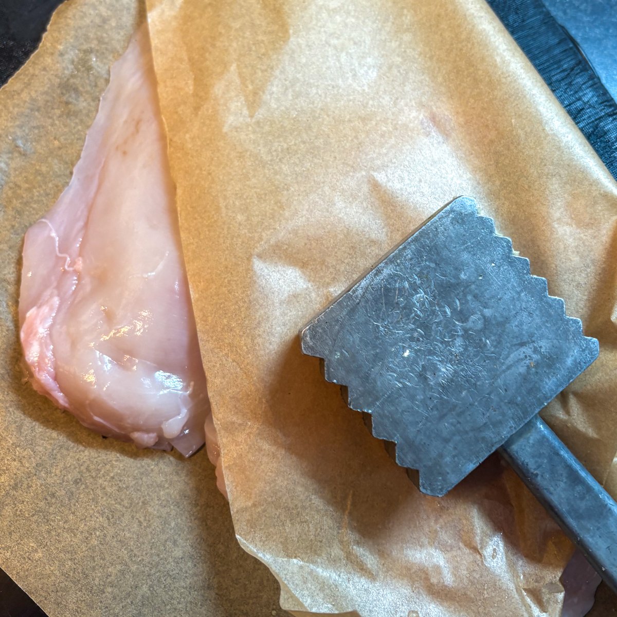 A raw chicken breast on parchment paper beside a metal meat hammer, ready to be flattened into even cutlets for pan-frying.