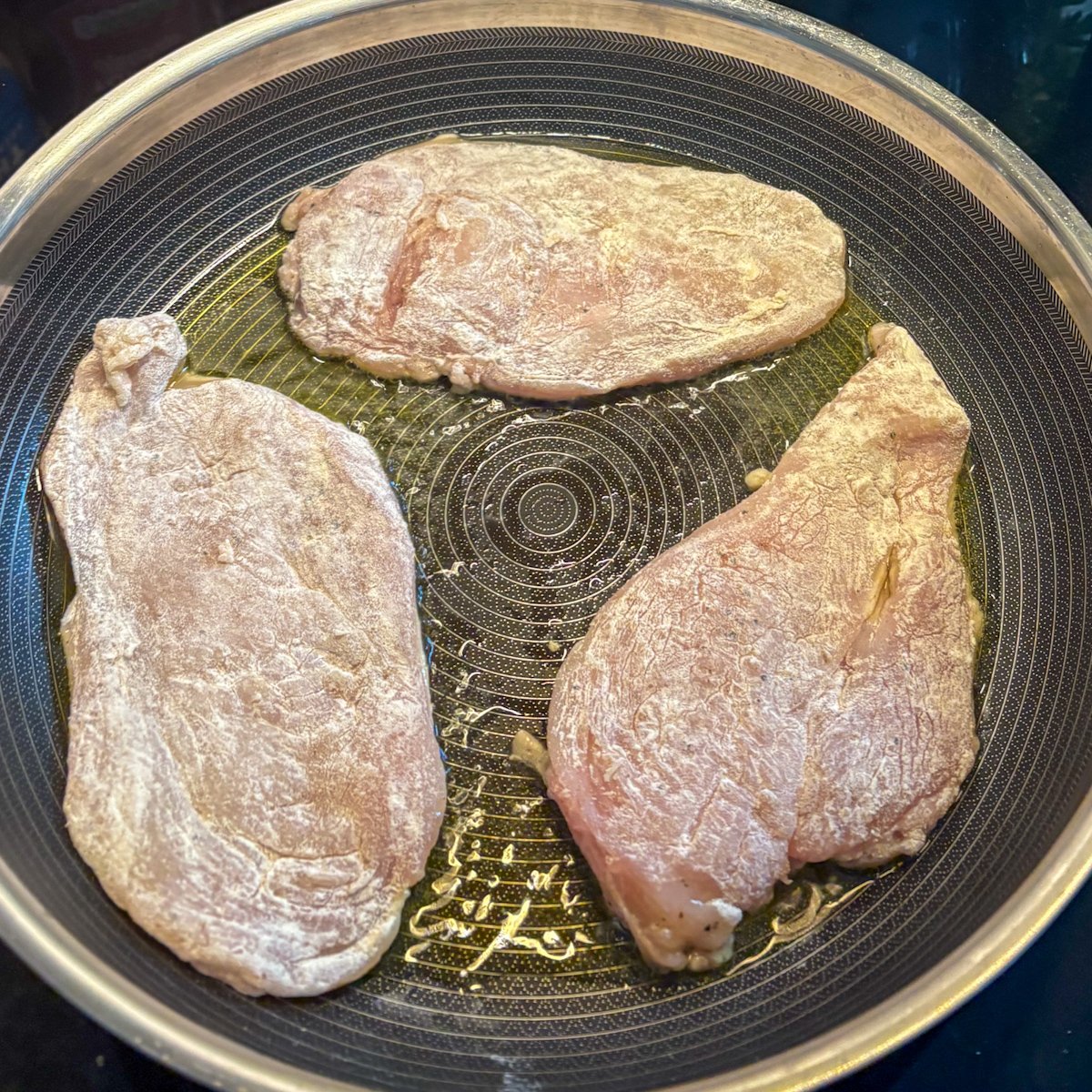 Three floured chicken cutlets frying in olive oil in a large non-stick pan, showing how to space them out so the pan is not overcrowded when making pan-fried chicken cutlets.
