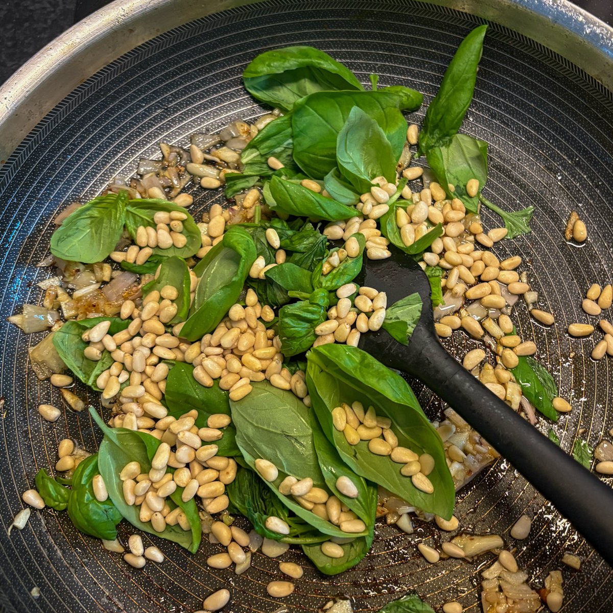 Fresh basil leaves, toasted pine nuts and chopped shallot being tossed in a frying pan with a spatula to make the topping for pan-fried chicken cutlets.